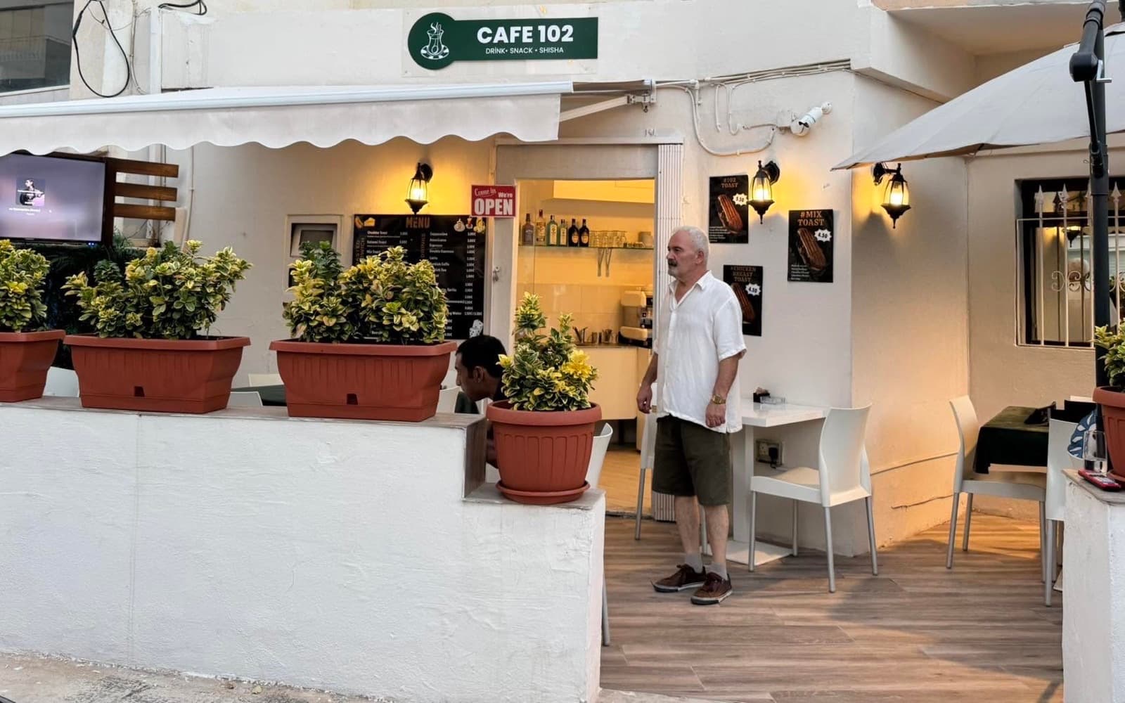 The Cafe 102 shopfront in Is-Swieqi — a green CAFE 102 sign above a white terrace with potted plants and outdoor tables, and the door open onto the counter.